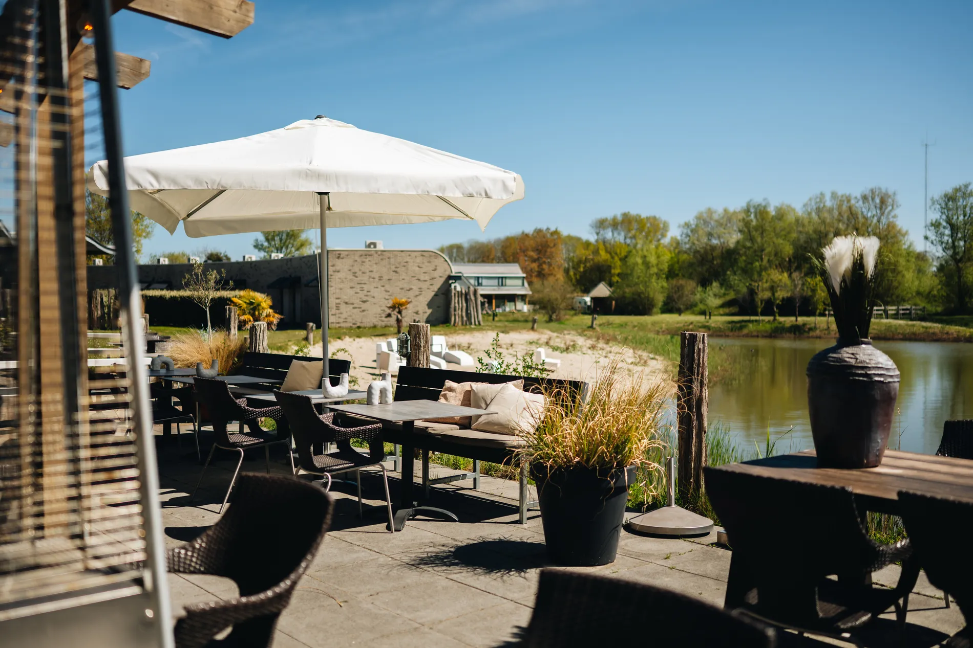 Overzicht van het terras met een grote witte parasol, comfortabele zitbanken en een weids uitzicht op een zandstrandje en de vijver van het resort.